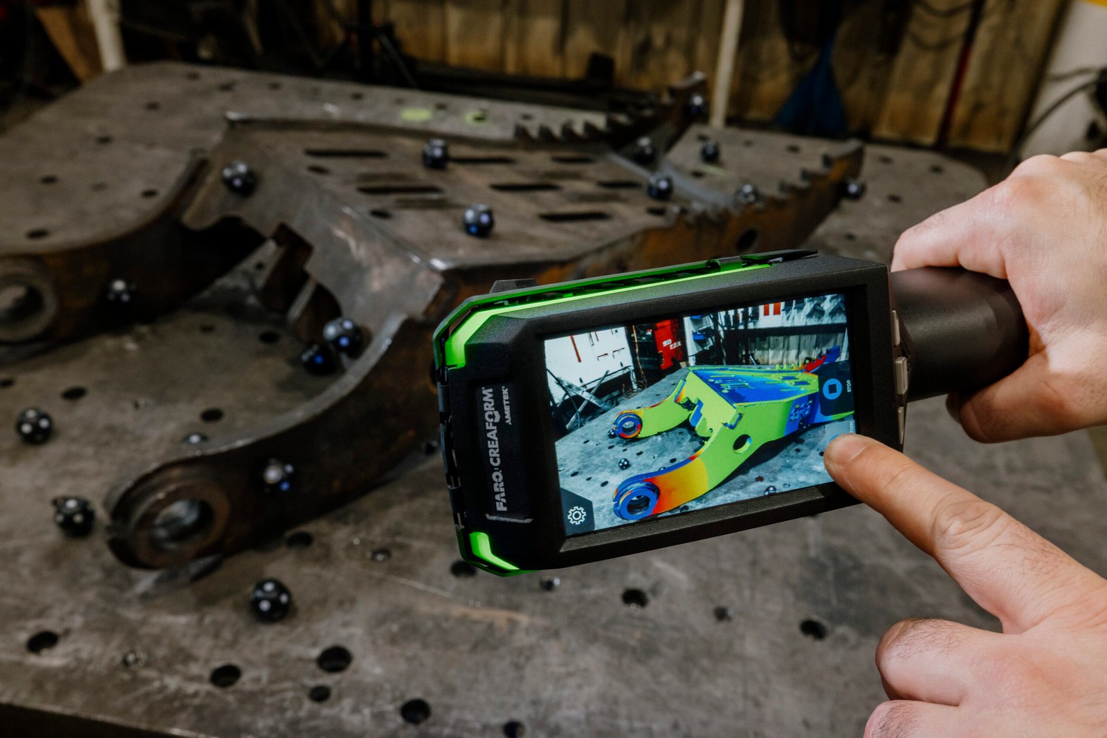 Handheld scanner displaying a colour-mapped digital view beside a large excavator component